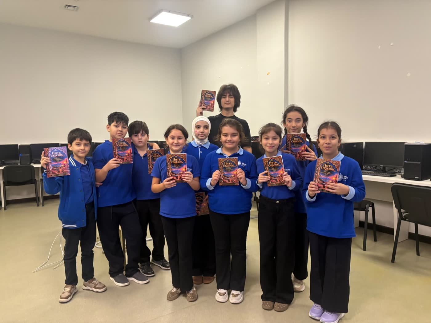 Nine pupils in blue uniforms stand in a computer room holding up the book, with Jamal behind them.