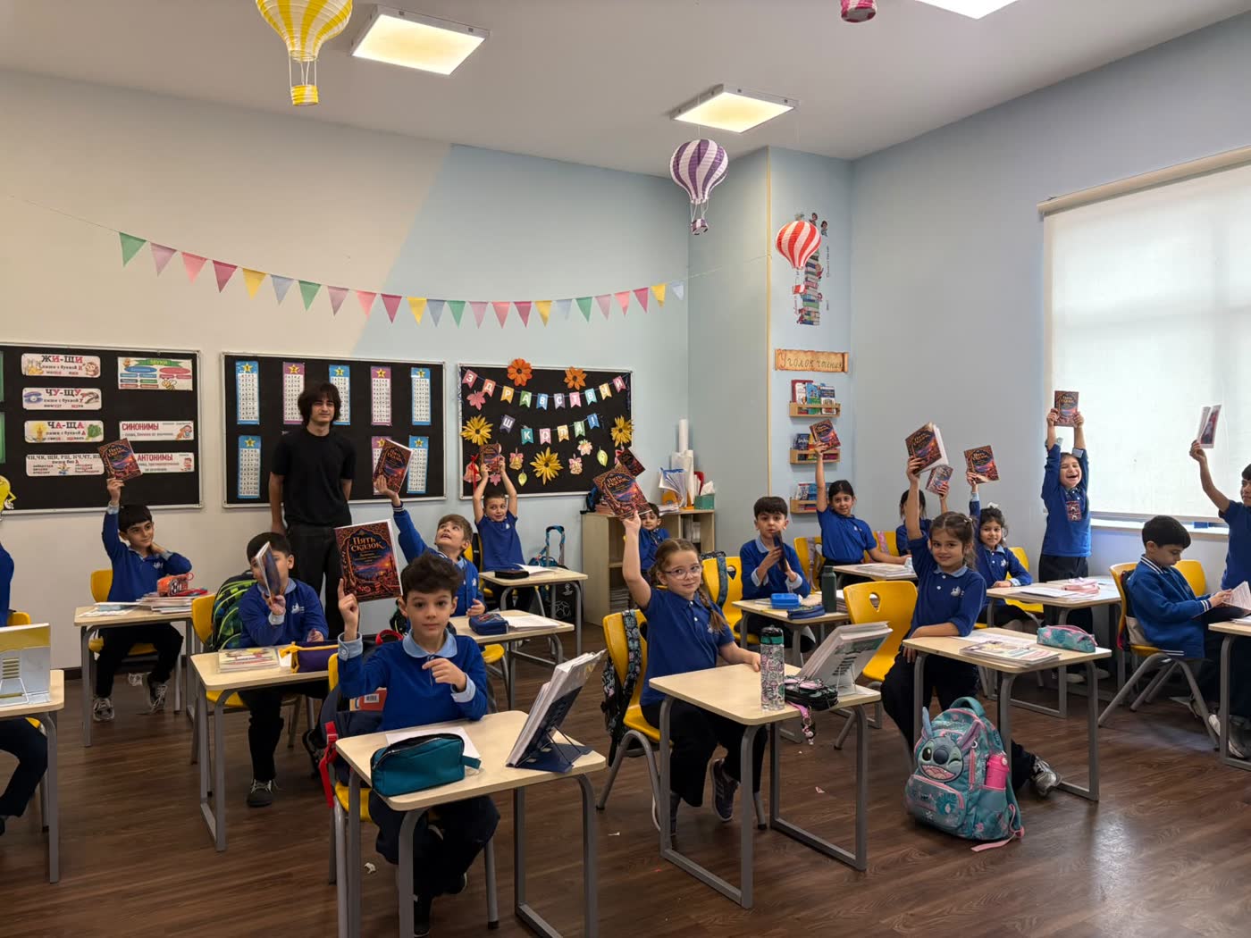 A classroom with paper bunting and hot-air balloon decorations; pupils hold the book above their heads.